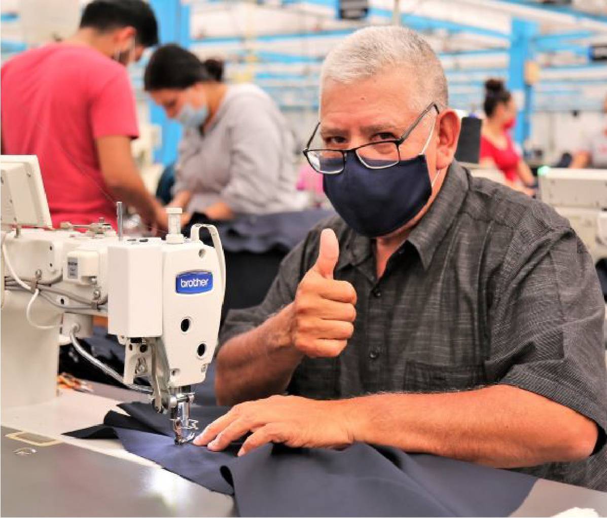 Male Industrias Manufactureras MyR seamstress wearing face mask holding his thumb up to camera