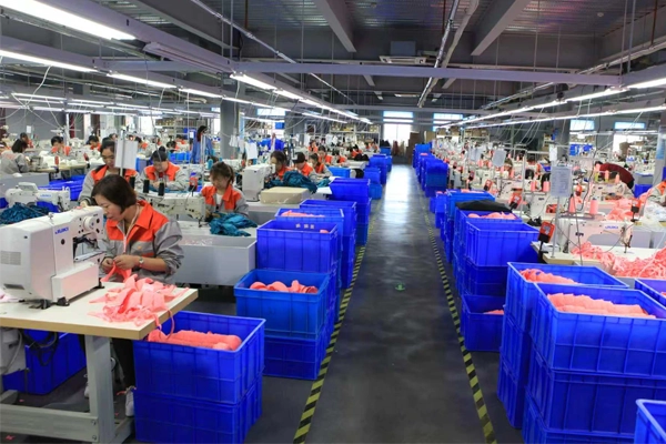 This image shows a large garment manufacturing facility with multiple rows of workers operating sewing machines. Blue storage bins filled with fabric pieces are arranged along a central aisle, and bright overhead lighting illuminates the organized production floor. The scene highlights a high-volume textile production environment focused on efficiency, quality control, and meeting customer demand.